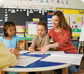 teacher sitting at desk with two students pointing at paper and
                    smiling